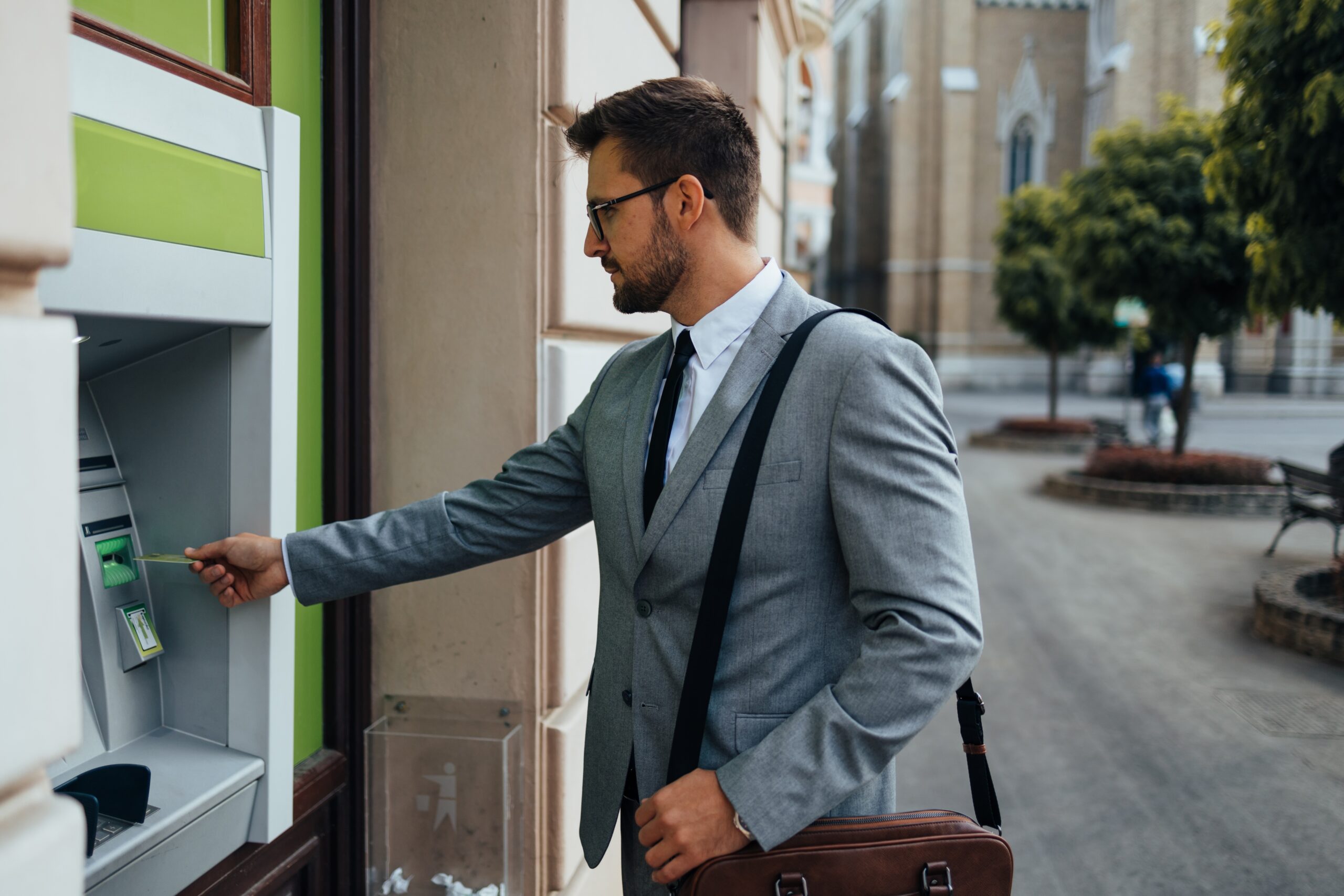 Businessman with eyeglasses standing on city street and using ATM machine to withdraw money from credit or debit card.